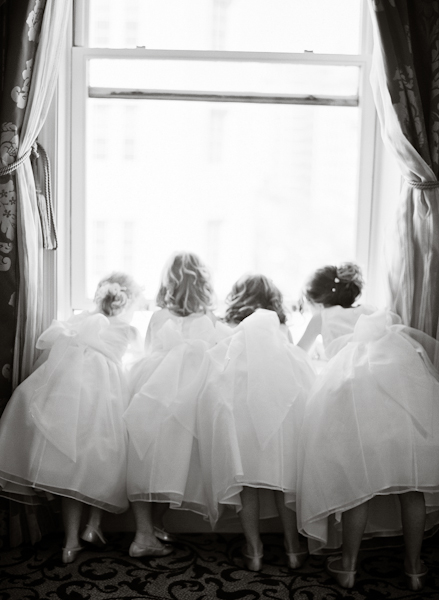 4 flower girls look out the window of the hotel suite while the bride gets dressed