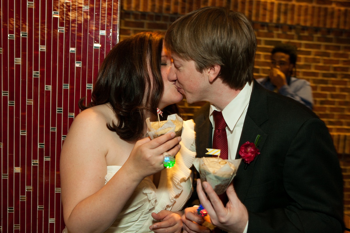 Bride and groom kiss while holding cupcakes from Baked & Wired