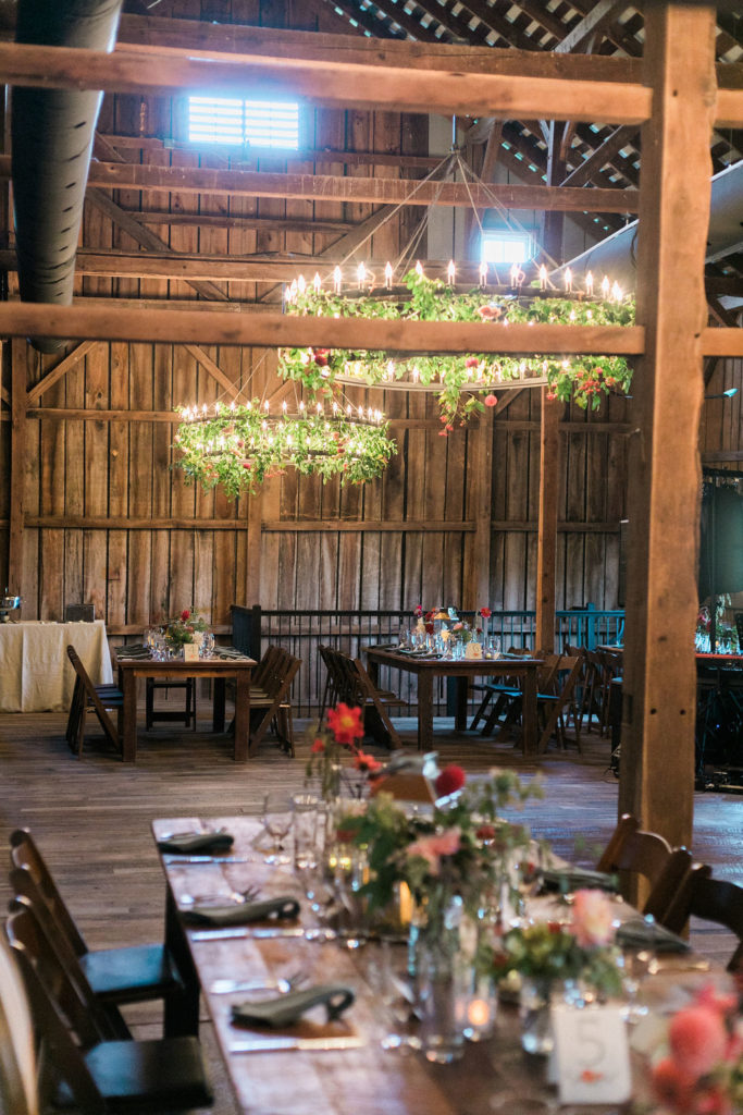 Tranquility Farm wedding reception in the barn  with farm tables and wooden folding chairs and florals and greenery adorning the chandeliers - planned by Bellwether Events